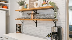 A kitchen with white cabinets, a coffee maker, and shelves holding glassware and decorative items. - Felspar House · Clean, Charming House in PB 🏖☀️ AC, pkg & laundry (San Diego)