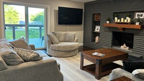 Una sala de estar con sofá, sillón, mesa de centro, chimenea y televisor montado en la pared.