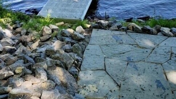 Un muelle de madera que se adentra en un lago, rodeado por un camino de piedra y una zona rocosa.