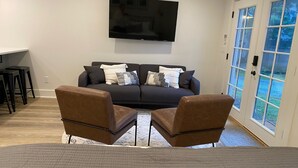 A modern living room with a dark sofa, two brown armchairs, a flat-screen TV on the wall, and a sliding glass door leading to a garden. - Remodeled Studio Cottage! Walk to Lake and Hiking Trails (Fall City)