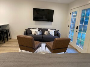A modern living room with a dark sofa, two brown armchairs, a flat-screen TV on the wall, and a sliding glass door leading to a garden.