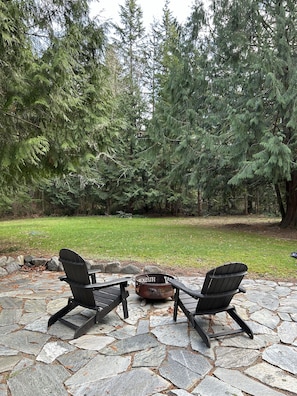 A stone patio with two Adirondack chairs facing a metal fire pit surrounded by a forest.