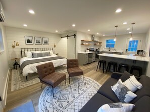 A modern bedroom with a large bed, a sitting area with a sofa and chairs, a kitchen with a bar, and a dining area.