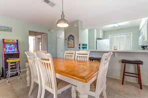 A dining area with a wooden table and white chairs, a kitchen area with a refrigerator, and a children's arcade game. - Bright & sunny 2 bed/2 bath condo in Navarre (Navarre)