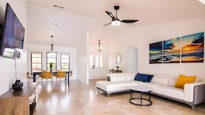 A modern living room with a white sofa, a dining area with yellow chairs, a flat-screen TV mounted on the wall, and a ceiling fan.