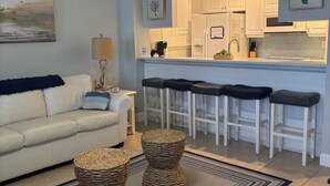 A modern living room with a white sofa, two wicker ottomans, a striped rug, and a ceiling fan. - RELAX and Save! Escape on sunny Fort Myers Beach in a 2BR/2BA w/ Pool/ Gulf view (Fort Myers Beach)