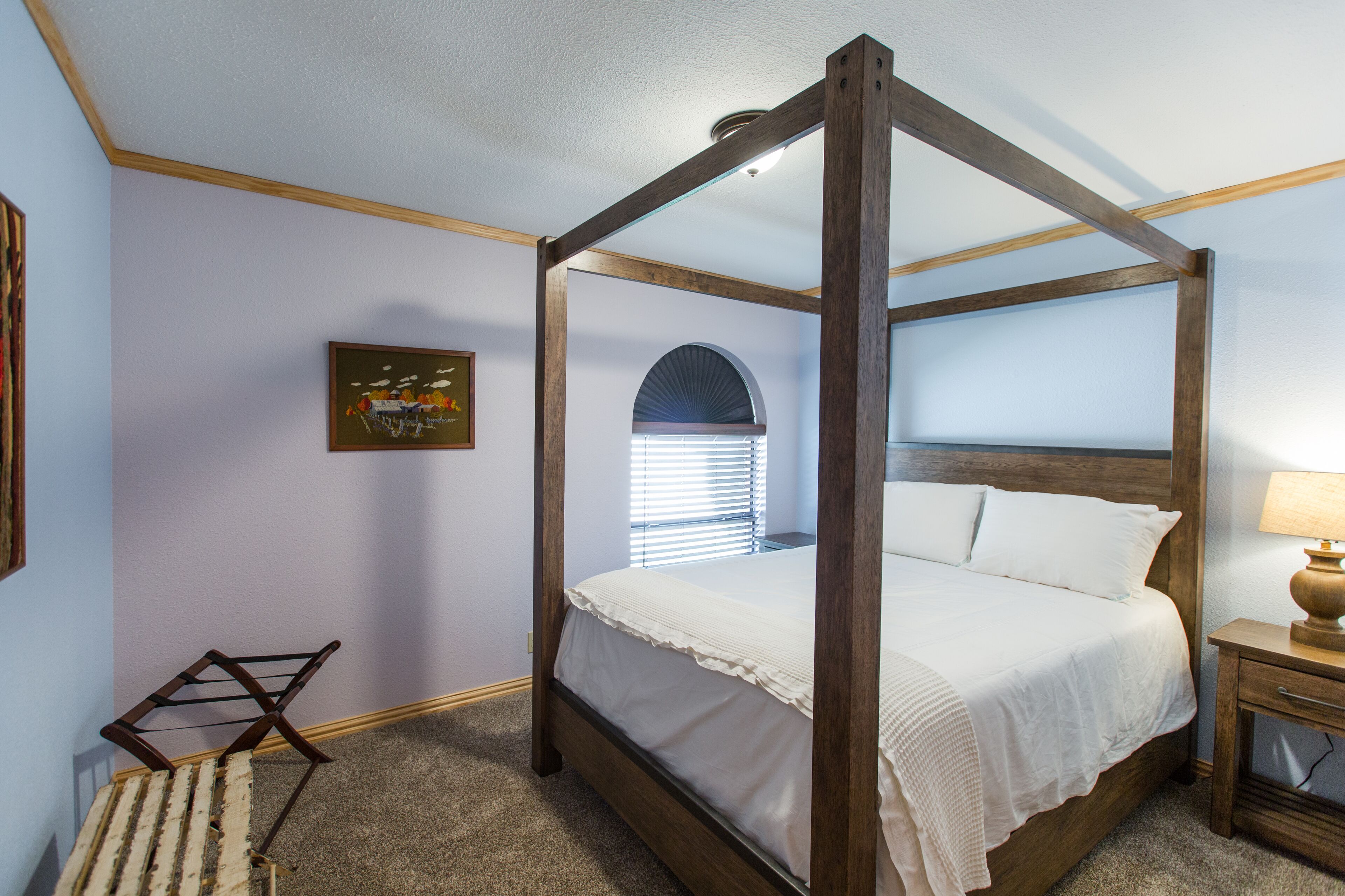 A four-poster bed with a wooden frame and white bedding, a small bedside table with a lamp, and a wall-mounted picture.