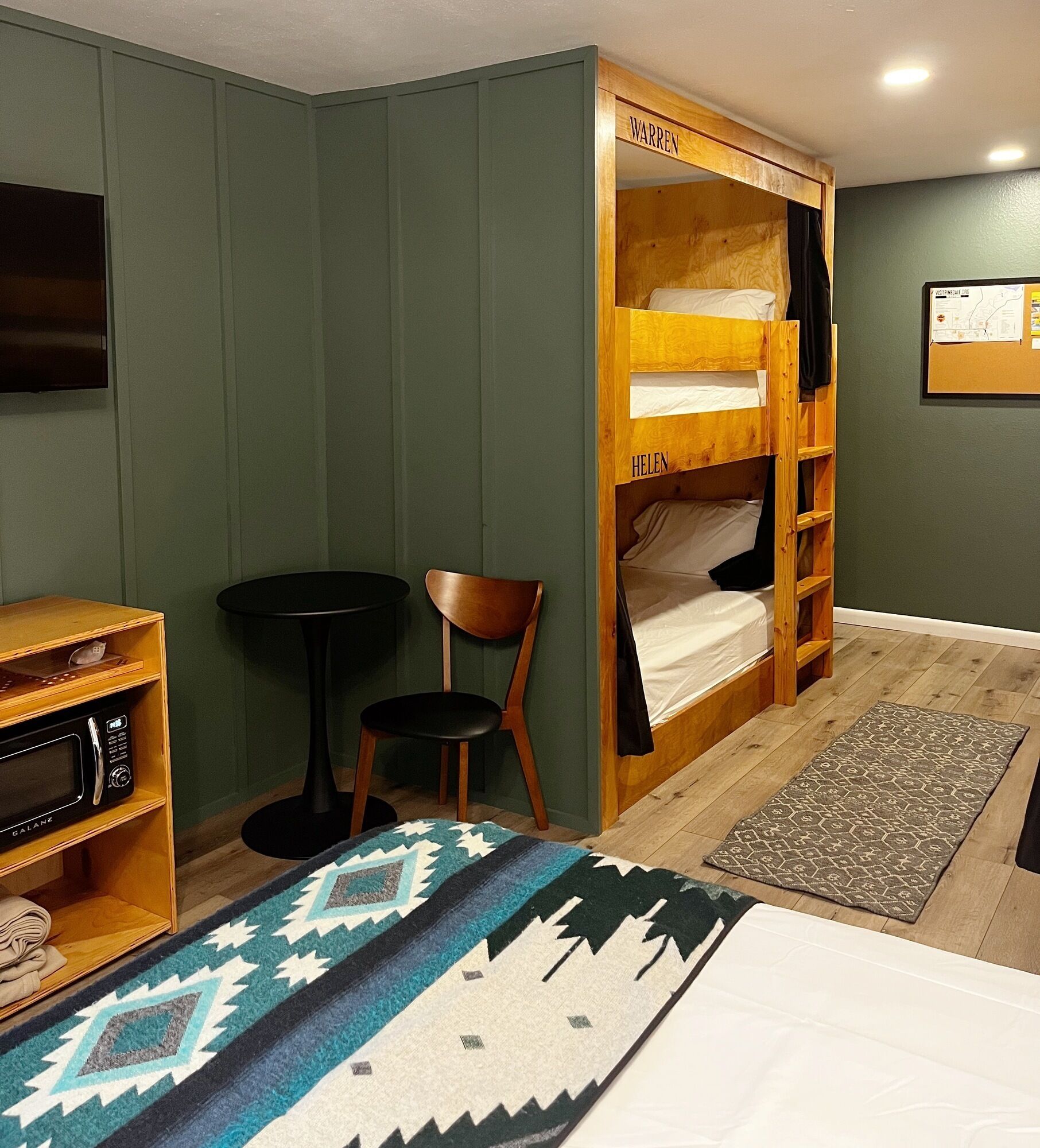 Quarto com beliche identificado como 'Warren' e 'Helen', uma mesa pequena, micro-ondas, uma cadeira e um tapete estampado.