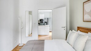 Ein modernes Schlafzimmer mit einem großen Bett, einem Nachttisch und einem kleinen Tisch daneben. Eine Tür führt in einen Küchenbereich mit Kühlschrank, Mikrowelle und Kaffeemaschine.