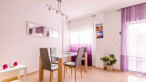 Un comedor con una mesa de vidrio, cuatro sillas, un banco blanco con jarrones rosados, una planta en maceta y una ventana con cortinas moradas.