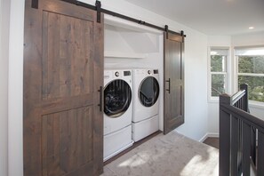 A laundry room with two front-loading washers and dryers, a sliding barn-style door, and a window with a view of trees. - Fully Renovated Condo in Greenspring Community (Mt Snow) (Dover)