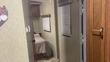 A narrow hallway with a bed, a door, and a window with blinds.