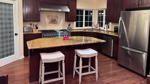 A kitchen with dark wooden cabinets, a stainless steel refrigerator, a granite countertop, and two white barstools. - Mountain Escape: Sleeps Multiple Families with Pool Table & Game Room (Running Springs)