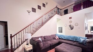 A living room with a wooden staircase, a sectional sofa, a small table with a lamp, and wall decorations. - Mountain Escape: Sleeps Multiple Families with Pool Table & Game Room (Running Springs)