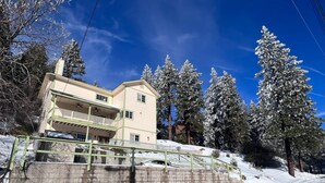 A two-story building with a balcony, surrounded by snow-covered trees and a clear sky. - Mountain Escape: Sleeps Multiple Families with Pool Table & Game Room (Running Springs)