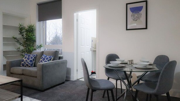 Een moderne woonkamer met een grijze bank, een glazen eettafel met vier stoelen, een klein bijzettafeltje en een potplant.