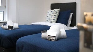 Hotelkamer met twee bedden, elk met een blauwe spreidek en witte kussens, waarvan één een blauw-wit patroon heeft. Er liggen handdoeken en aan de linkerkant staat een klein tafeltje met een lamp.