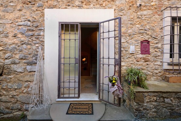 Un edificio de piedra con una puerta abierta, un felpudo, una planta en maceta y una ventana pequeña con un buzón.