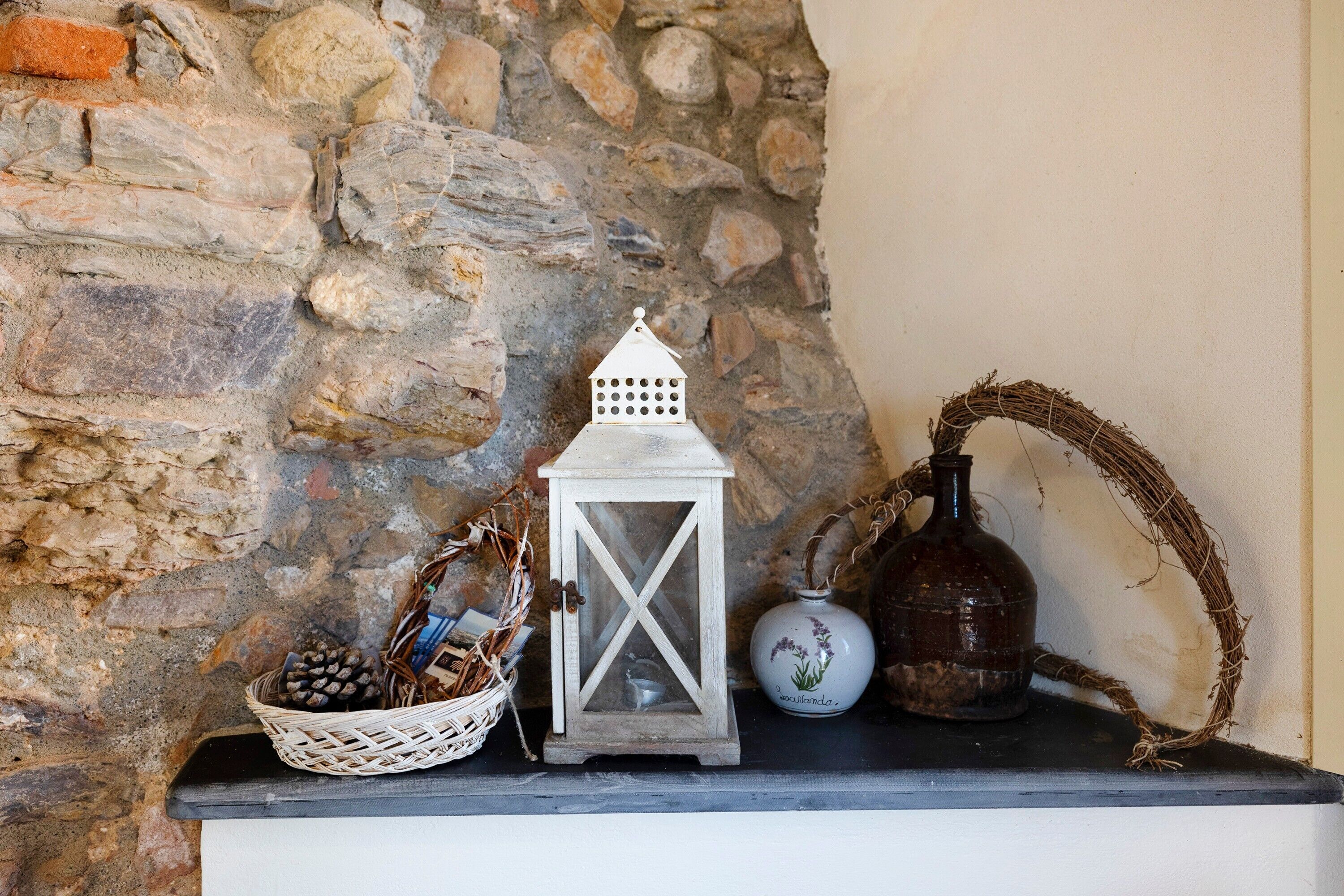 Un mur de pierres, une lanterne blanche, un panier en osier rempli de pommes de pin et un vase de fleurs séchées posé sur une étagère.