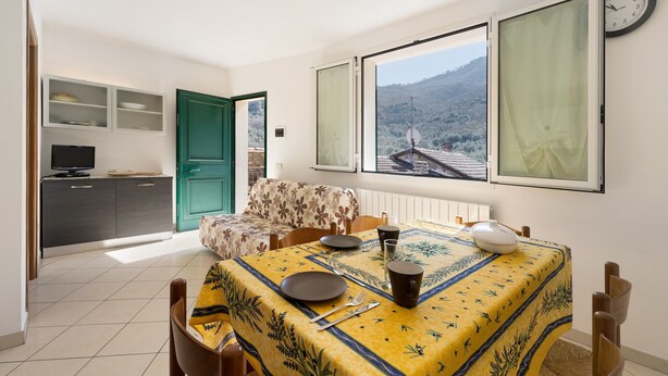 A dining area with a table set for two, a sofa, a television, and a view of the outdoors. - Apartment 'Pana La Taggiasca' with Mountain View (Stellanello)