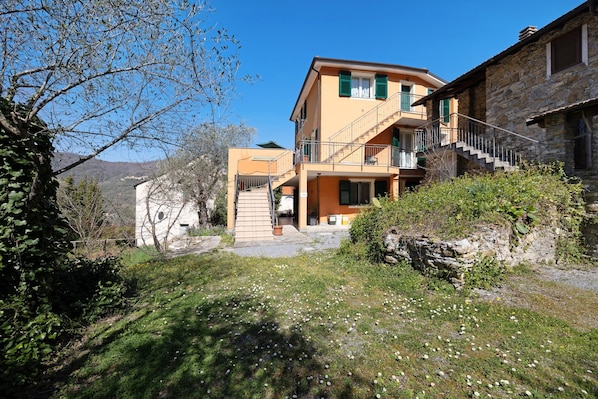 A two-story building with a staircase leading to a balcony, surrounded by greenery and a clear sky.