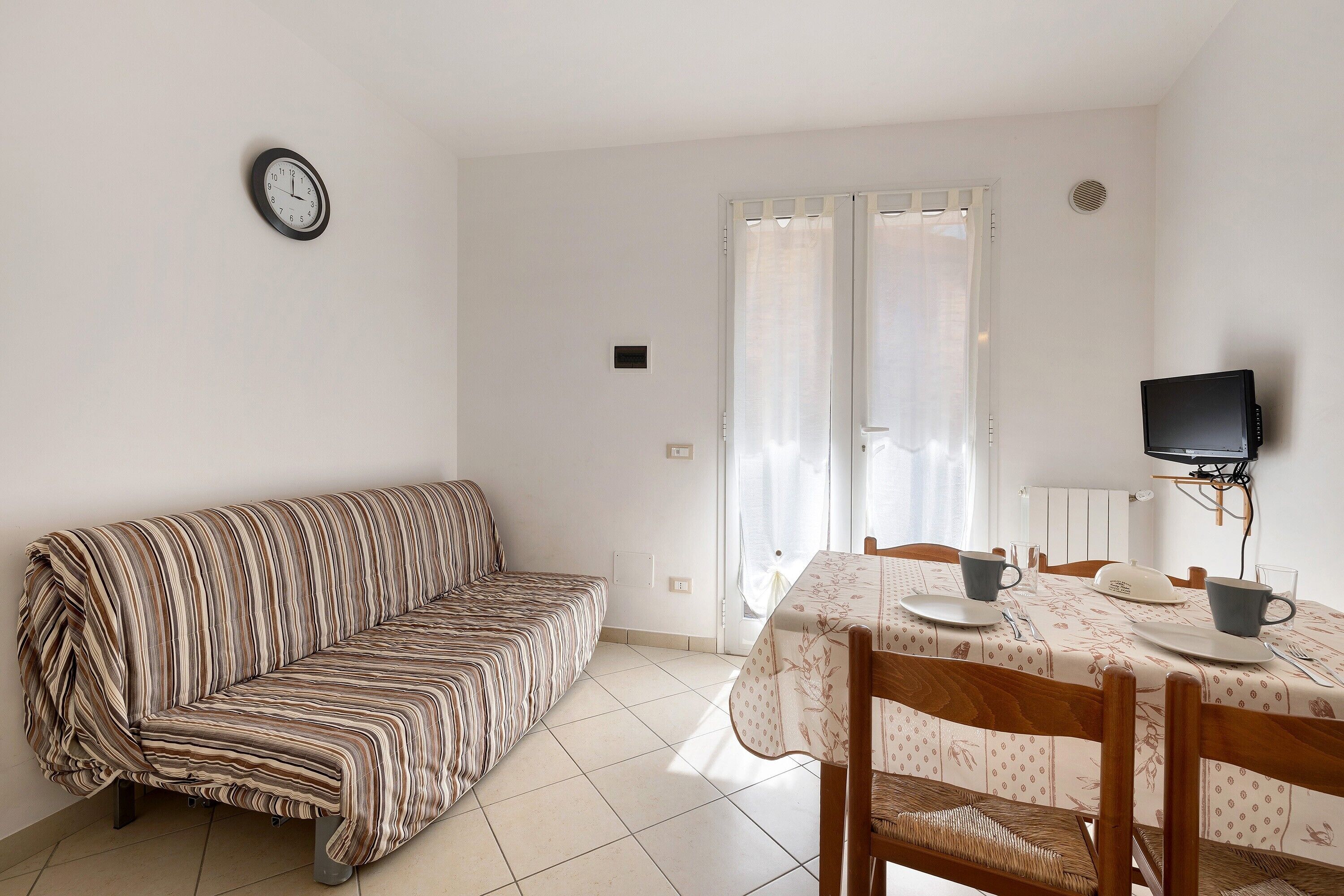 A room with a striped sofa, a dining table with chairs, a TV, and a window with curtains.