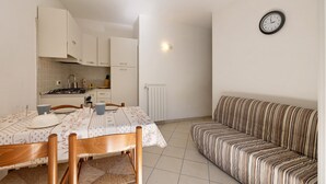 Een kleine keuken met witte kastjes, een eettafel met stoelen, een gestreepte bank en een wandklok.