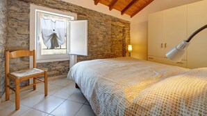 Een slaapkamer met een stenen muur, houten plafond, een bed met een bedsprei met patroon, een houten stoel, een raam met gordijnen en een bedlampje.