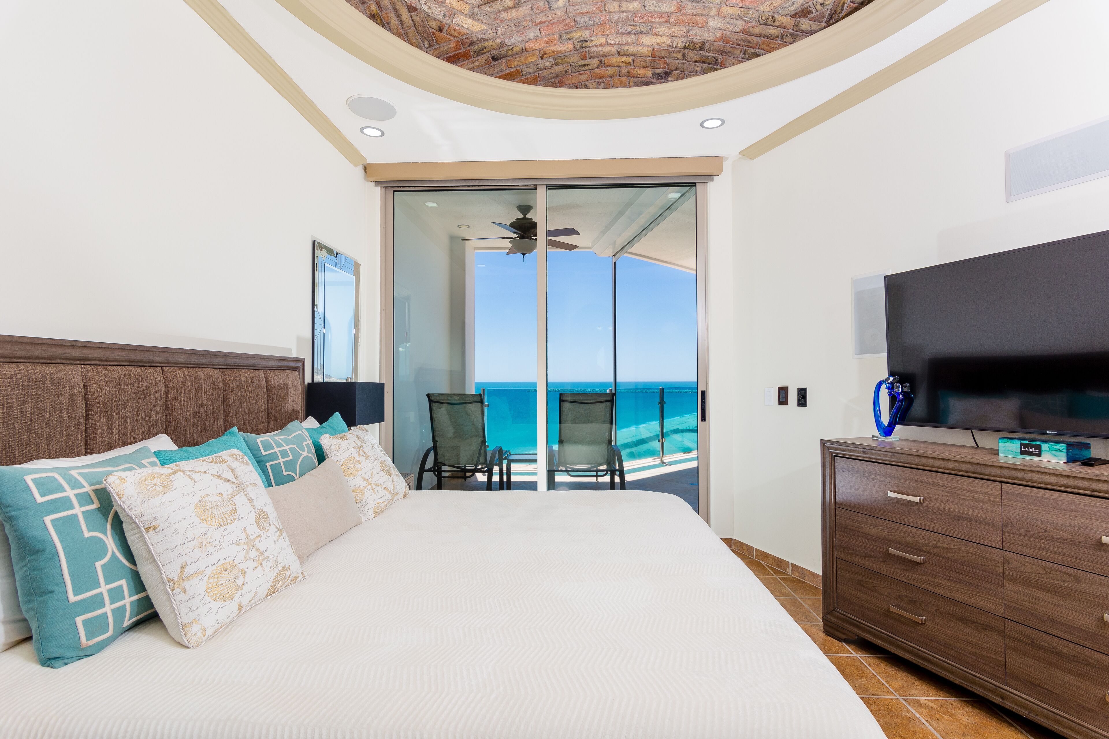 A bedroom with a large bed, a TV, a dresser, and a balcony with a view of the ocean.