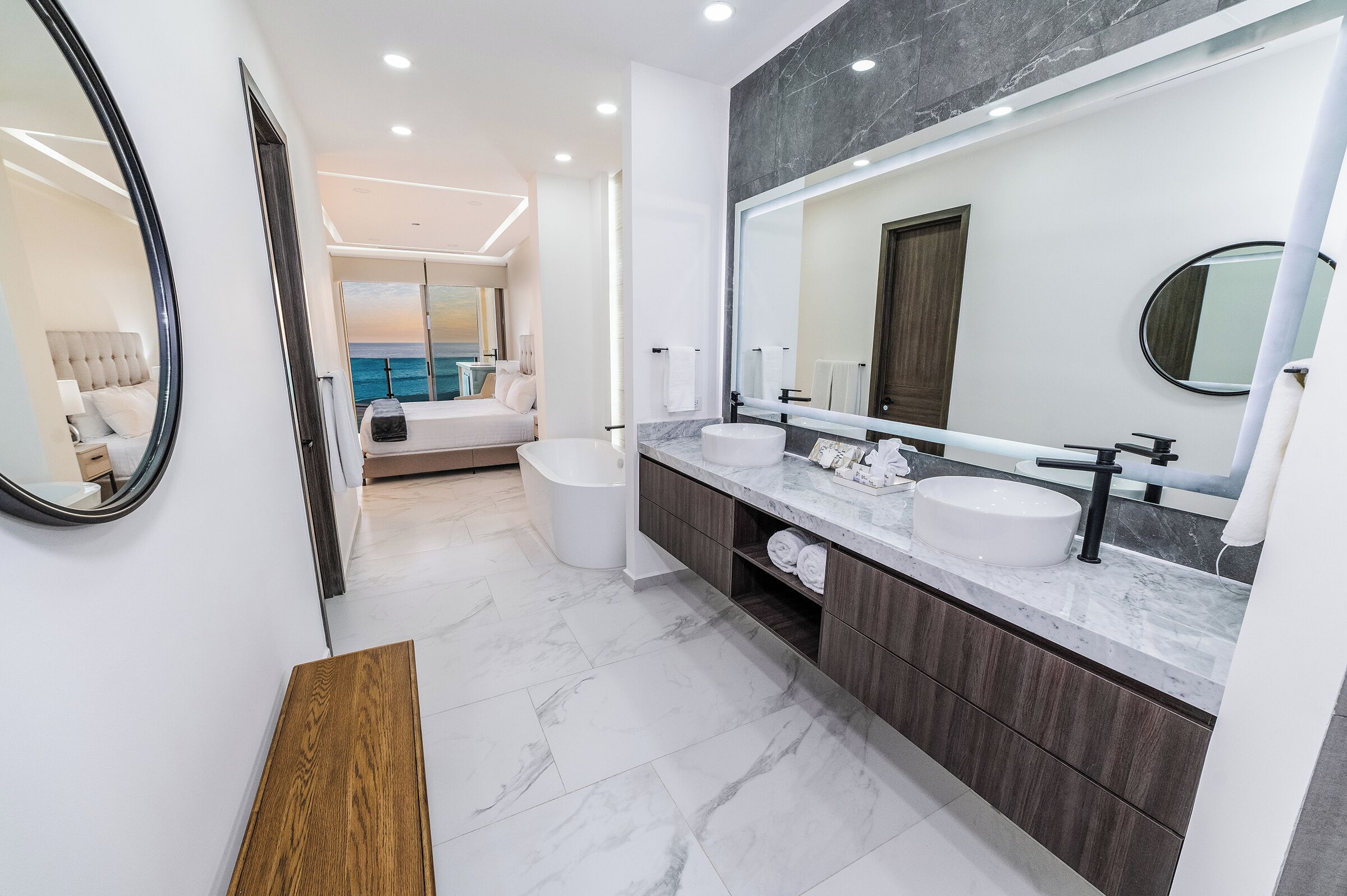 Een moderne badkamer met dubbele wastafels, een grote spiegel en uitzicht op een slaapkamer met een bed en uitzicht op zee.
