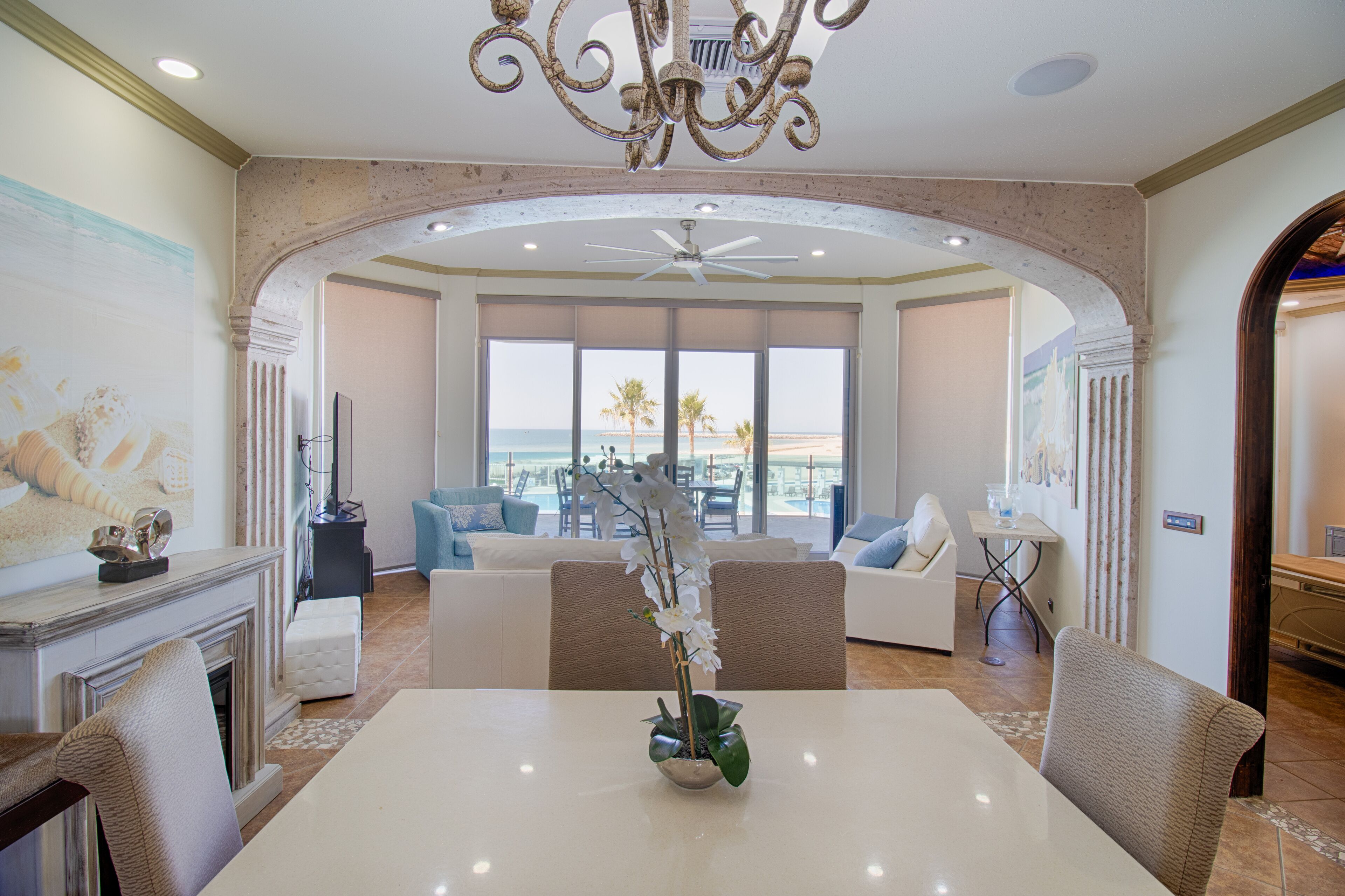 A spacious living room with a large window overlooking a beach, a fireplace, and a dining area with a white table and chairs.