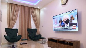 A living room with a TV mounted on the wall displaying a news broadcast, two green chairs, a wooden coffee table, and a wooden cabinet.