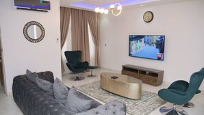 A modern living room with a grey tufted sofa, teal armchairs, a wooden coffee table, and a flat-screen TV mounted on the wall.