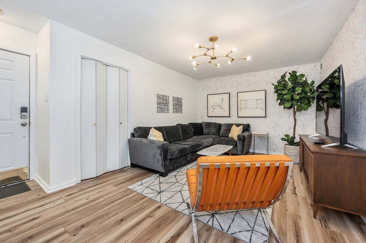 Een moderne woonkamer met een grijze hoekbank, een oranje fauteuil, een houten salontafel en een tv op een houten dressoir.