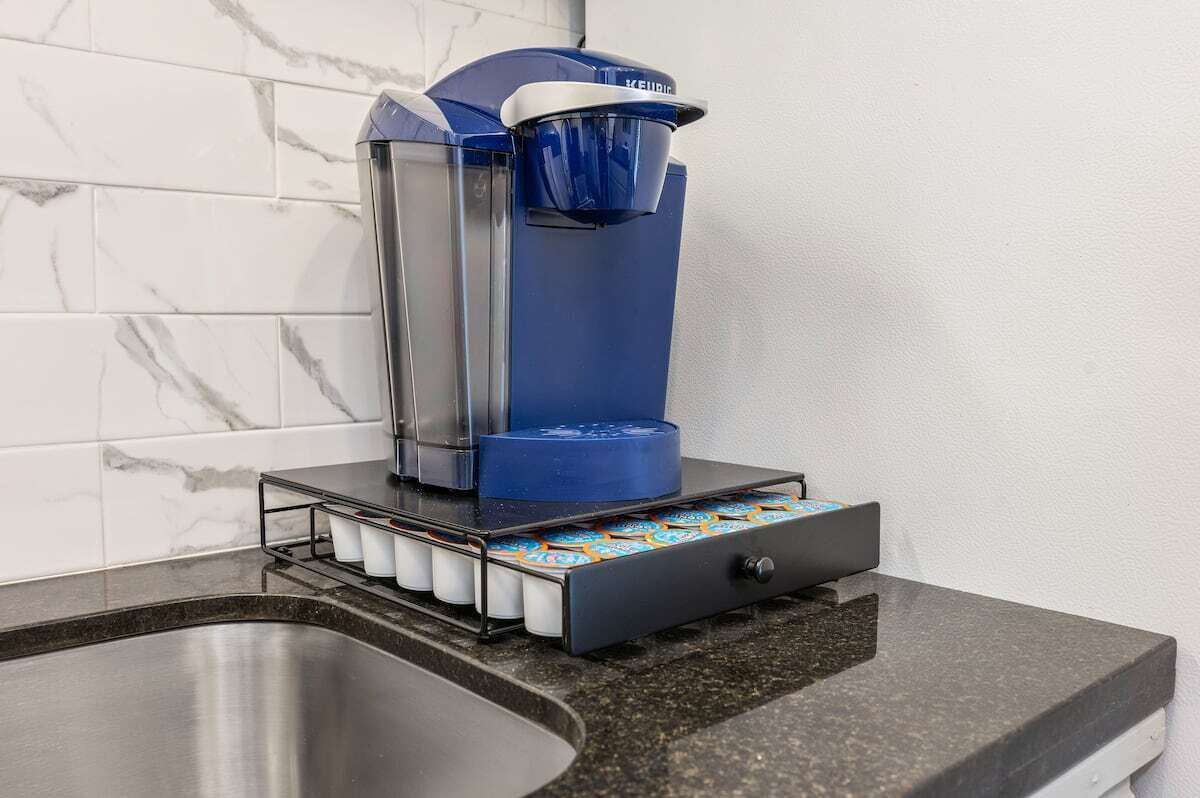 Eine Kaffeemaschine mit blauem Sockel und einem transparenten Wasserbehälter, auf einer schwarzen Arbeitsplatte neben einem Spülbecken platziert.