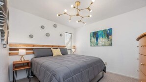 Quarto com cama, mesas de cabeceira, cômoda, luminária de teto e um quadro na parede.