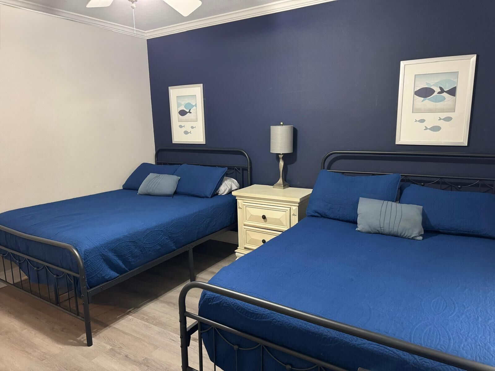 Een kamer met twee eenpersoonsbedden, elk met een blauwe sprei en bijpassende kussens. Tussen de bedden staat een nachtkastje met een lamp. De muren zijn donkerblauw en er hangen afbeeldingen in lijsten.