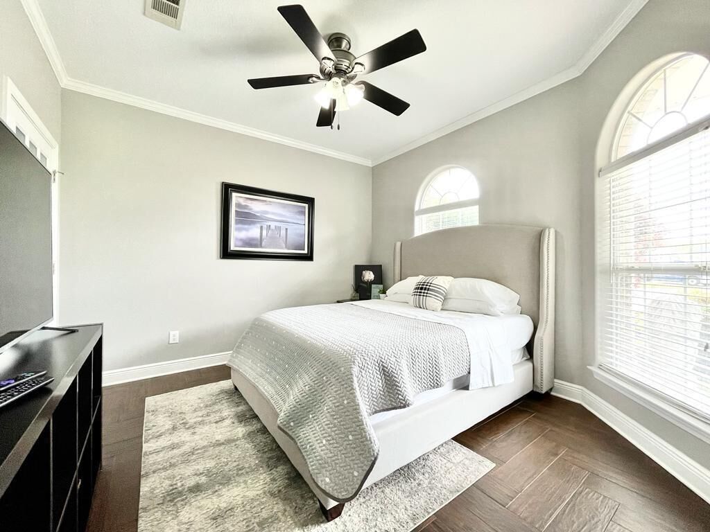 A bedroom with a bed, a ceiling fan, a window with blinds, a framed picture on the wall, and a desk with a computer.