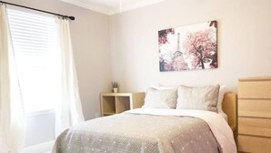 Een slaapkamer met een bed, nachtkastje, dressoir en een raam met gordijnen.