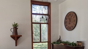 A living room with a piano, a sofa, a coffee table, a wooden shelf with a plant, and a large wall clock. - Rustic modern cabin surrounded by trees and large yard 25 miles from Waco/Temple (Lott)