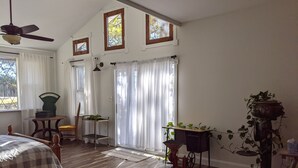 A bedroom with a bed, a ceiling fan, a window with curtains, a desk with a chair, and a potted plant. - Rustic modern cabin surrounded by trees and large yard 25 miles from Waco/Temple (Lott)