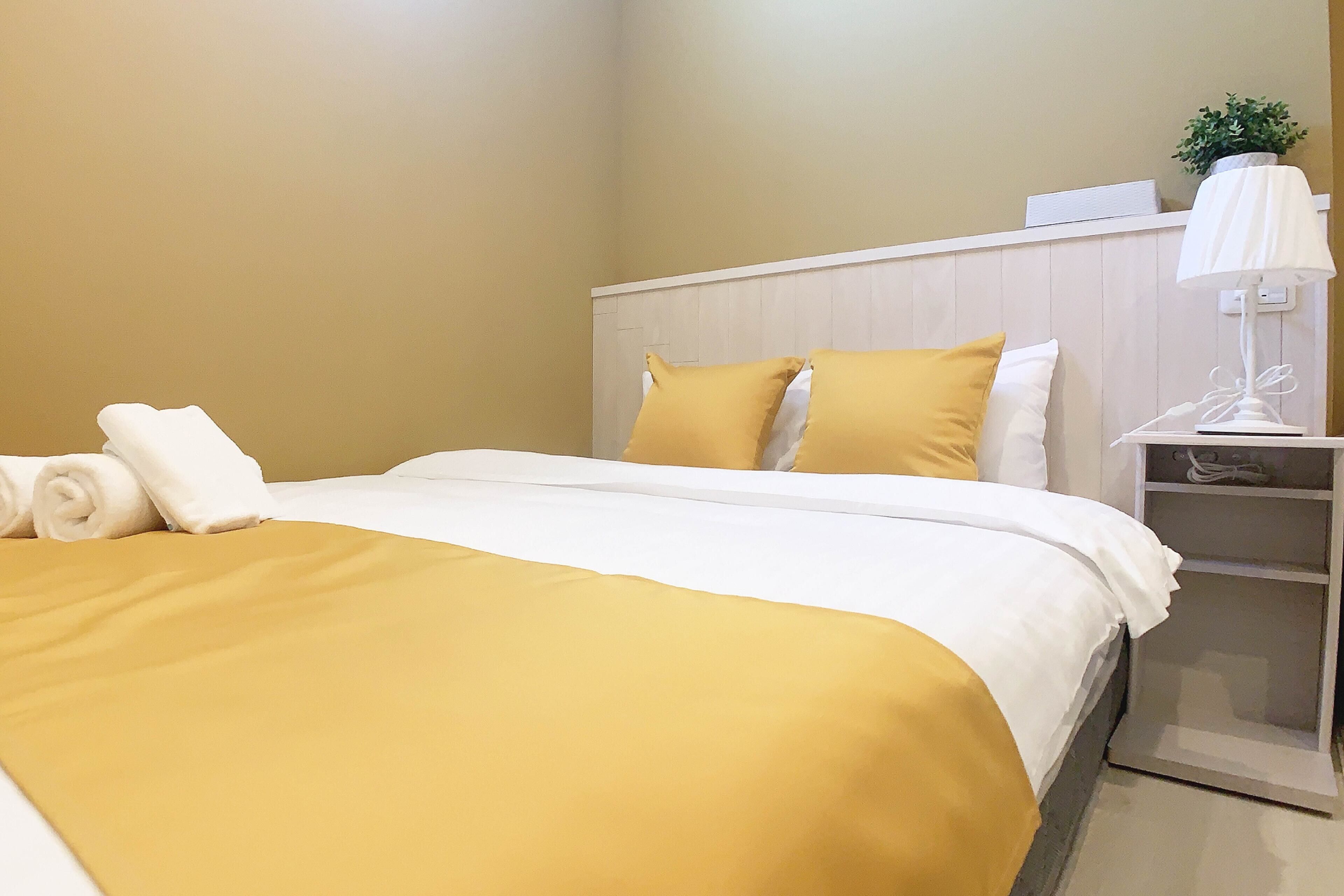 Una cama bien hecha con una colcha amarilla y blanca, una almohada blanca y una toalla blanca enrollada a un lado.