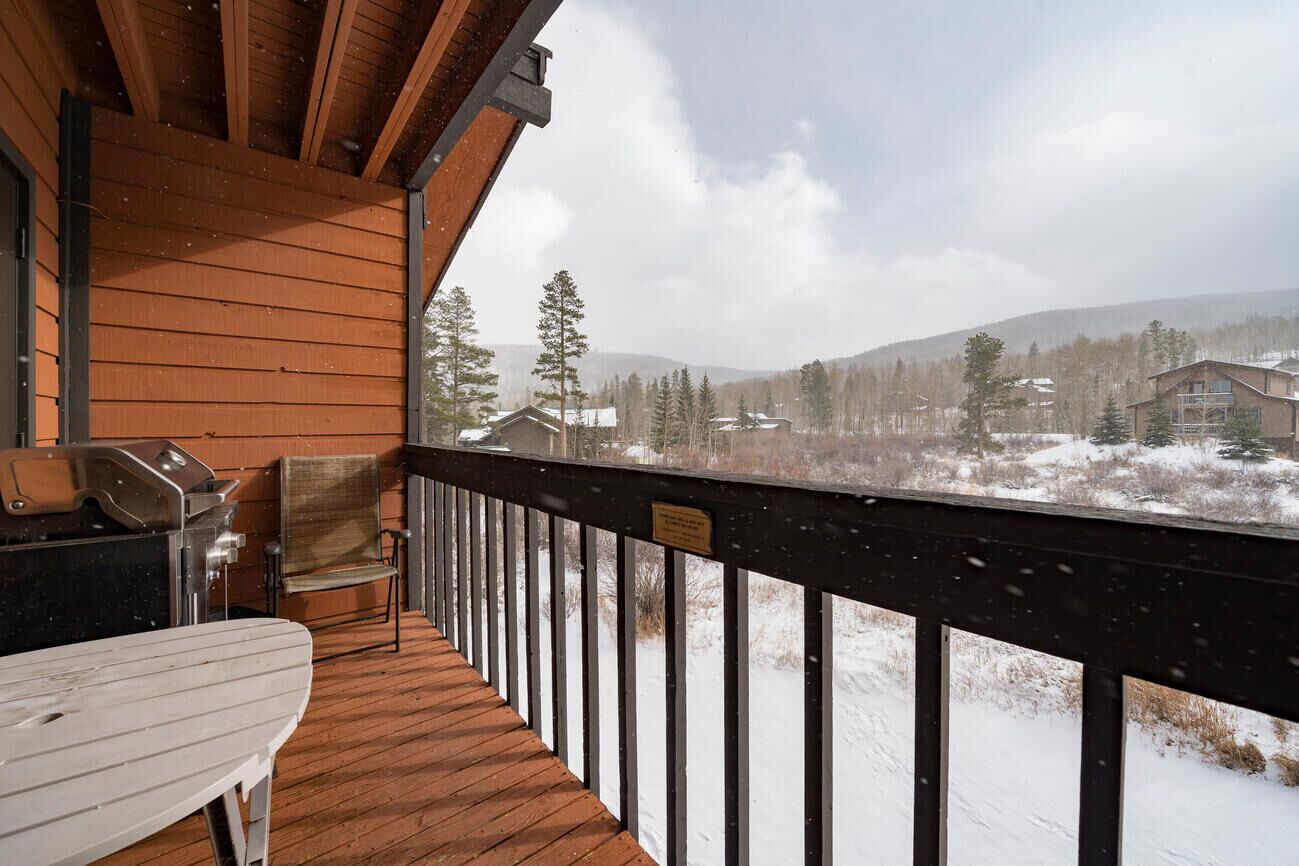 A wooden deck with a grill and a table, overlooking a snowy landscape with trees and buildings.