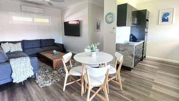 Un salon moderne comprenant un canapé, une table basse et une table à manger avec des chaises. Un téléviseur est fixé au mur, il y a un ventilateur de plafond et un coin cuisine avec des placards et un plan de travail.