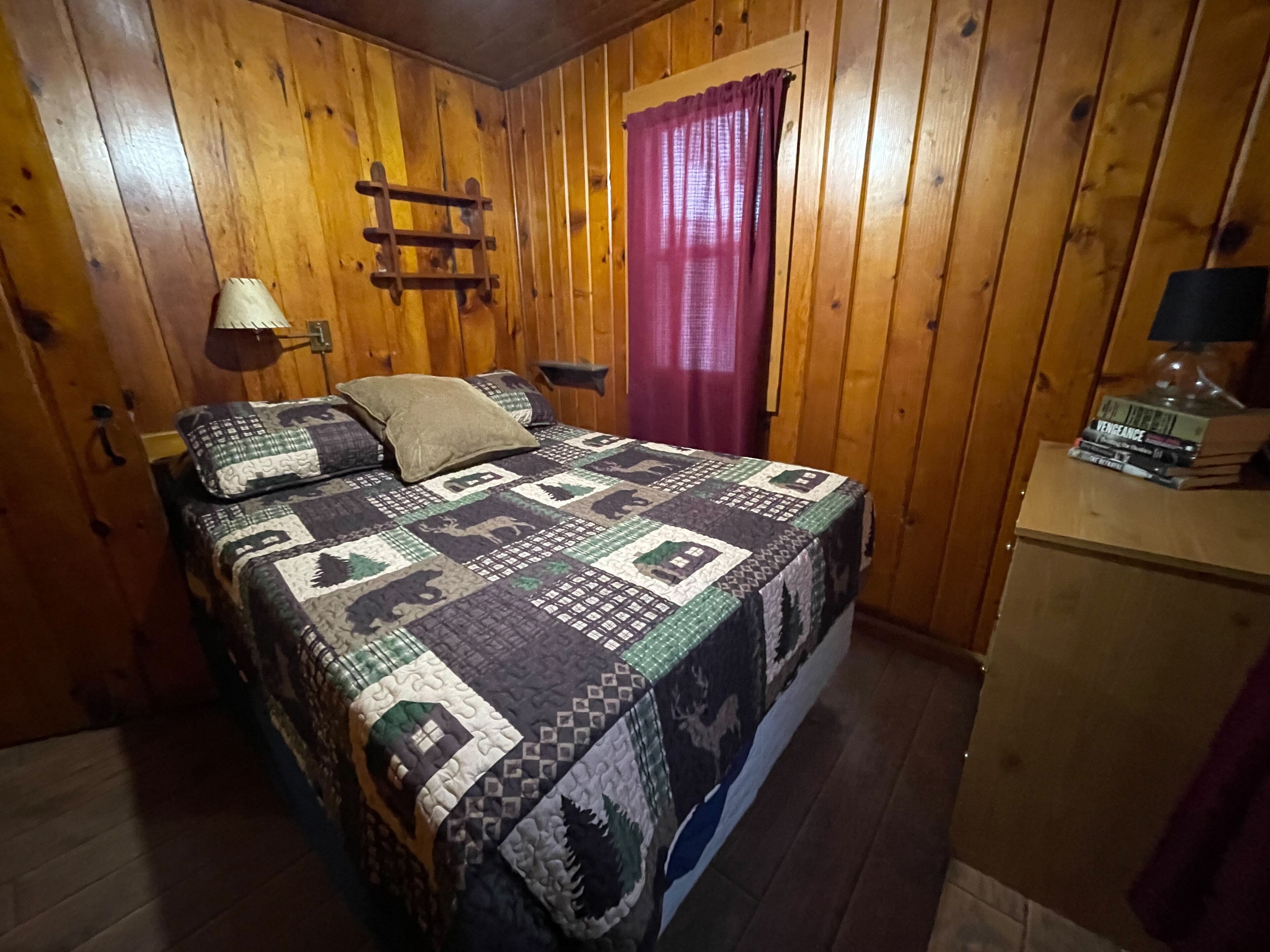 A wooden-paneled room with a bed, a nightstand, a lamp, and a window with curtains.