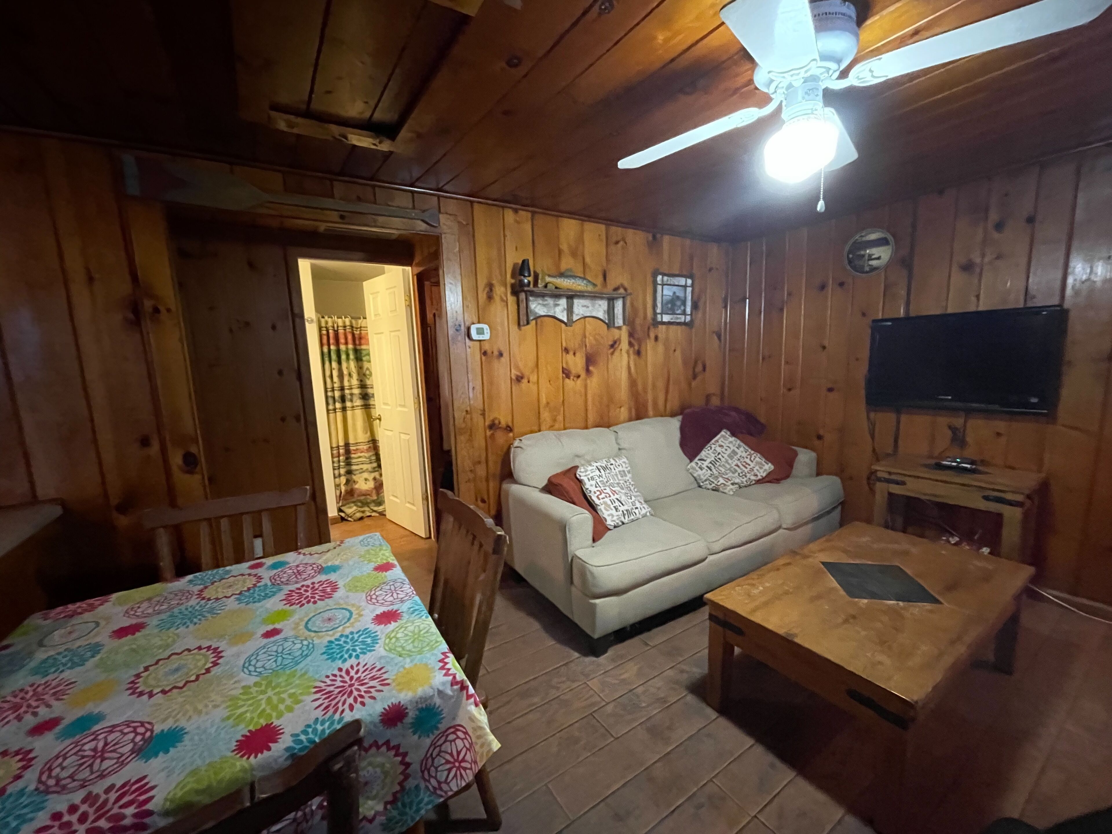 Une chambre confortable, aménagée dans le style d’un chalet, avec un plafond en bois, un ventilateur de plafond, une télévision, un canapé agrémenté de coussins à motifs, une table basse en bois et une table recouverte d’une nappe colorée.
