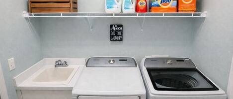 A laundry room with a sink, two washing machines, and a shelf with laundry supplies.