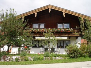 A traditional wooden chalet with a balcony adorned with flowers, a stone pathway leading to the entrance, and outdoor seating. - Ferienwohnung für 5 Personen, 2 Schlafzimmer, 70 m² (Inzell)