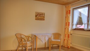 A room with a table and two wicker chairs, a radiator, and a window with curtains. - Ferienwohnung für 5 Personen, 2 Schlafzimmer, 70 m² (Inzell)