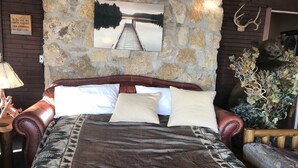 A bed with a brown blanket, a headboard, and pillows. There is a stone wall behind the bed with a picture of a lake and a wooden bench to the right. - NEW-Waterfront Lodge-Private dock/slip, Kayak, swim, fish, water view from deck (Grove)